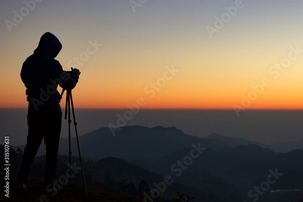 Fototapeta silhouette of Travel photographer standing with a camera mounted on a tripod and shooting a time lapse of the sunrise/sunset. man wearing his hood enjoying the mountain view /valley view