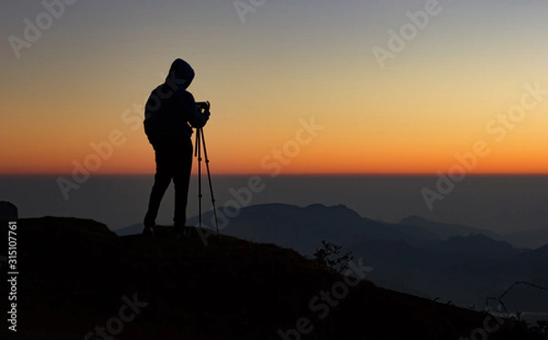 Fototapeta silhouette of Travel photographer standing with a camera mounted on a tripod and shooting a time lapse of the sunrise/sunset. man wearing his hood enjoying the mountain view /valley view