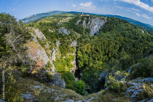 Fototapeta Unesco world heritage site Skocjanske jame taken with a wide fish eye view. Looking towards the deep gorge with cave entrance and walking paths. Village of Skocjan in the background.