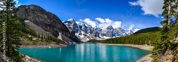 Fototapeta This pristine Moraine Lake overlooks the icy rocky mountains and pine forest. The light breeze gently ripples the turquoise water towards the rocky edge on this partially cloudy day.