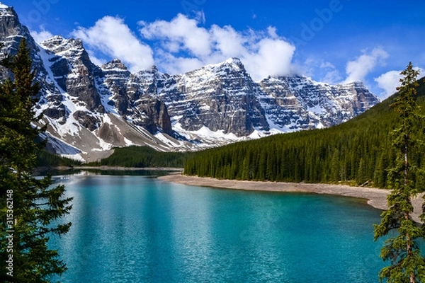 Fototapeta This pristine Moraine Lake overlooks the icy rocky mountains and pine forest. The light breeze gently ripples the turquoise water towards the rocky edge on this partially cloudy day.