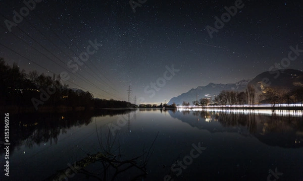 Fototapeta Night landscape by a lake, reflection of the sky in the lake, lights of vehicle headlights on the highway