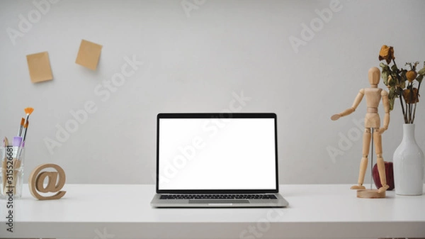 Fototapeta Close up view of  workspace with blank screen laptop, painting tools, wooden figure and decorations on white desk with white wall