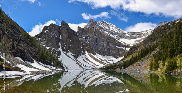 Fototapeta The mirror like reflections on Lake Agnes create beautiful patterns against the mountainous backdrop spotted with snow and pine trees on a sunny and partially cloudy day.