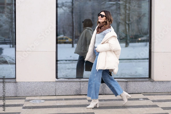 Fototapeta European model girl in a beige oversized down jacket, knitted sweater, flared jeans with a handbag and glasses is walking down the street. Life style