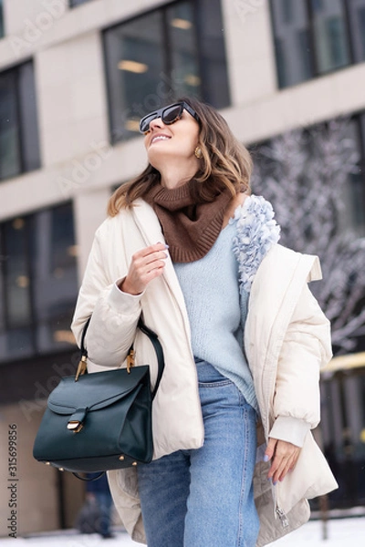 Fototapeta Caucasian model girl in a beige oversized down jacket, knitted sweater with a handbag and glasses posing near the office building. Lifestyle. Real emotions