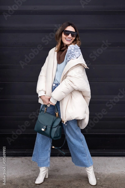 Fototapeta Caucasian girl in oversized down jacket, knitted sweater, flared jeans with a handbag and glasses posing on the street. Life style