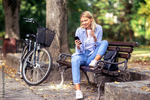 Fototapeta Blonde young woman sitting on the bench looking on mobile phone and drinking coffee to go in park with bicycle on the background