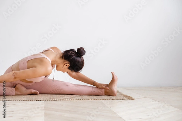 Obraz Close-up of a pacified young well-groomed brunette woman doing stretching while doing Pilates exercises sitting on a rug in a pink gymnastic suit on a white background. Place for advertising