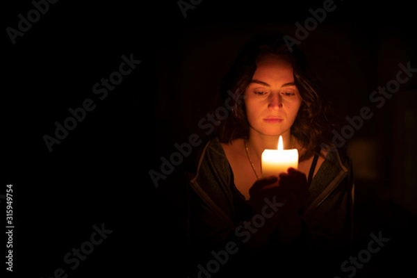 Fototapeta A sad religious orthodox brunette girl with brown eyes looks piercing and sad looking, holding a bright, lighted candle in folded hands. The girl on the right. Horizontal view