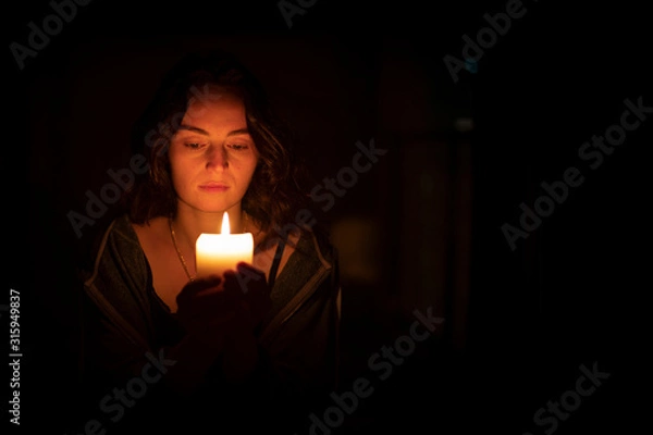 Obraz A sad religious orthodox brunette girl with brown eyes looks piercing and sad looking, holding a bright, lighted candle in folded hands. The girl on the left. Horizontal view