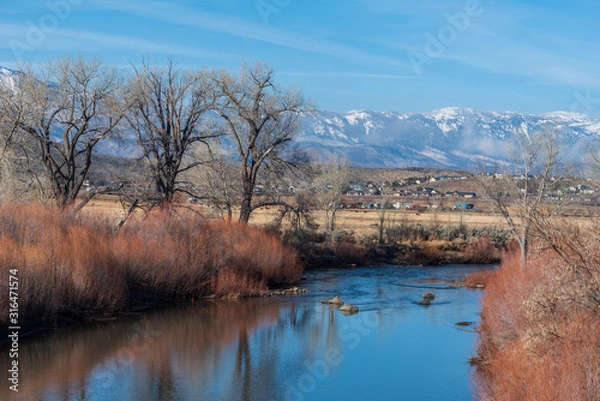 Fototapeta Landscape of the Carson River, bare trees, red reeds and snow-dappled mountains in the distance in Carson City Nevada
