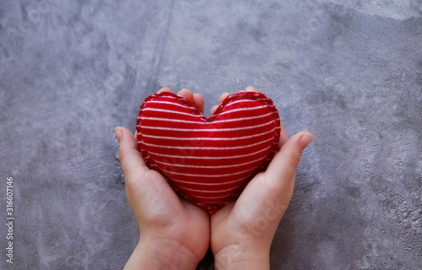 Obraz Textile soft red heart with white stripes in the hands on a gray concrete background.Selective focus.Cotton heart for Valentine's day, hand-sewn. A gift for February 14, made with your own hands
