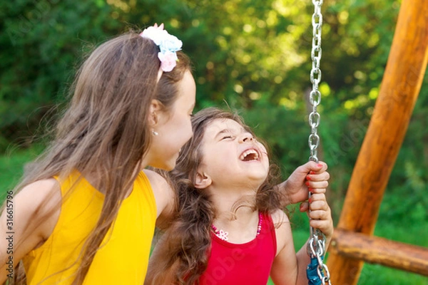 Obraz Two happy beautiful girls sitting on swing and smiling at warm  summer day