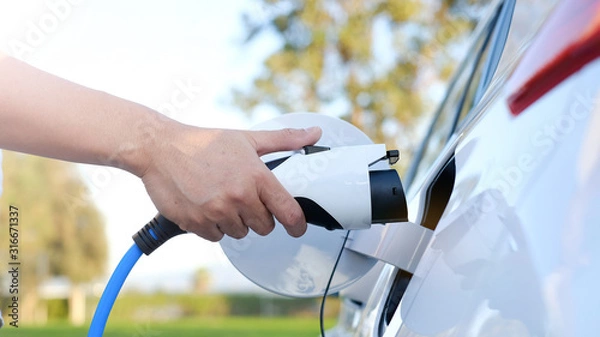 Fototapeta Electric car or ev is charging at station . man use the white power cable and plug on nature background. eco and clean energy concept with the mobile phone smart technology. zero gas no transmission