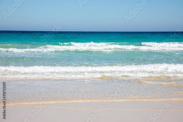 Fototapeta Cloudless sky over a colorful sea with diferent colors aquamarine turquoise dakr blue azure with waves prducing white foam crushing against a yellow beach summer sumertime sunny calm water tranquil