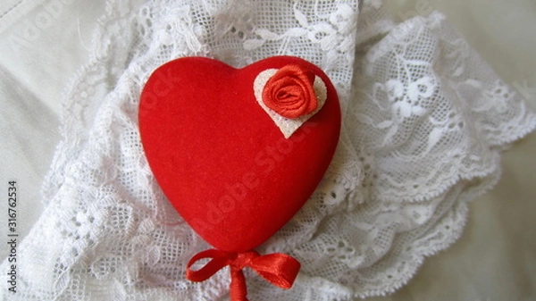 Fototapeta A red heart on a stick with a small red bow, decorated with a small red rose, lying on white lace panties. Saint Valentine's Day gift concept. 