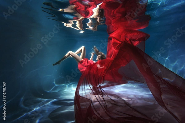 Obraz Underwater scene. A woman, a fashion model in the water in a beautiful dress swims like a fish.