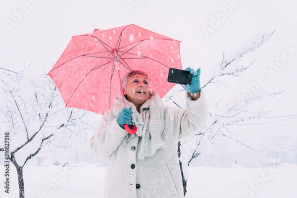 Obraz older woman with red umbrella taking a picture with lots of snow 