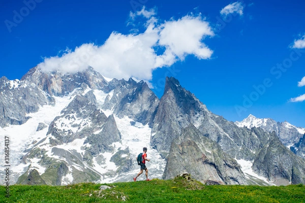 Fototapeta A trail runner running below the high snowy mountains along a grassy ridge in the Alps along the Tour du Mont Blanc route.