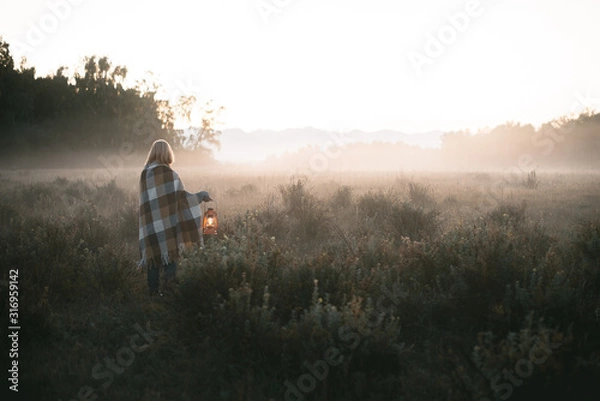 Fototapeta young girl with blond hair in a warm woolen sweater wrapped in a plaid, she is standing in a field with tall dry grass at dawn with a kerasin lamp in her hands