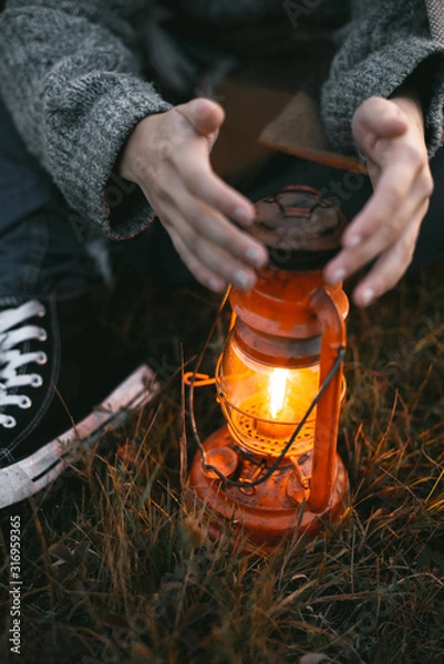 Fototapeta vertical shot of a young girl with blond hair in a warm woolen sweater wrapped in a plaid, she sits in a field with tall dry grass at dawn with a kerasin lamp
