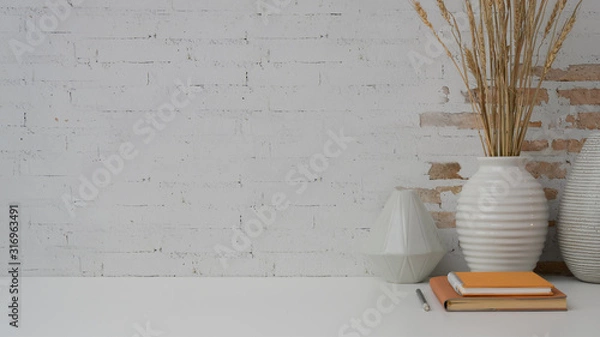 Fototapeta Close up view of workplace with copy space, notebooks and ceramic vase on white desk with brick wall