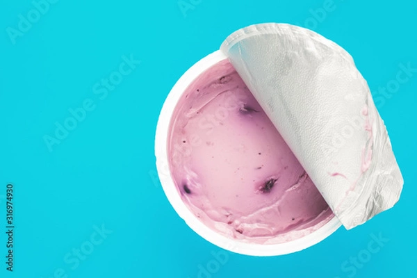 Fototapeta A pink yogurt with blueberry in an open plastic cup on blue background. Top view, text space.