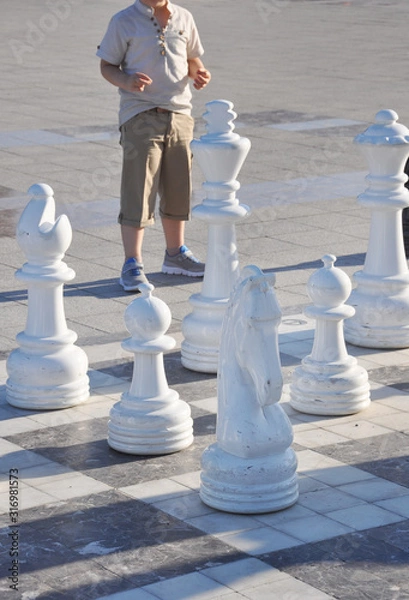 Fototapeta Boy plays giant chess on the street. White chess pieces on a chessboard lined with colorful concrete tiles on the sidewalk