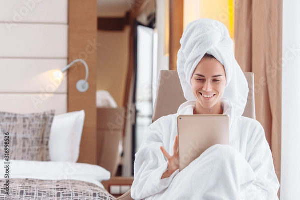 Obraz Entrepreneurial positive young business woman relaxing in spa hotel on business trip filling out an activity diary using a tablet while sitting on a couch in a bathrobe and a towel on her head