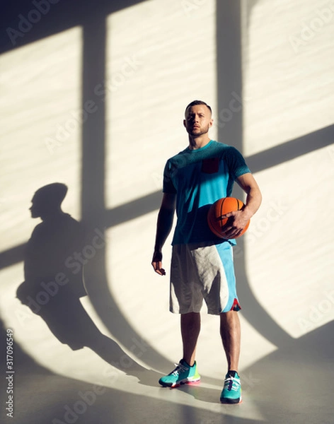 Obraz Young athletic man, basketball player holding ball and standing near wall with shadows from window