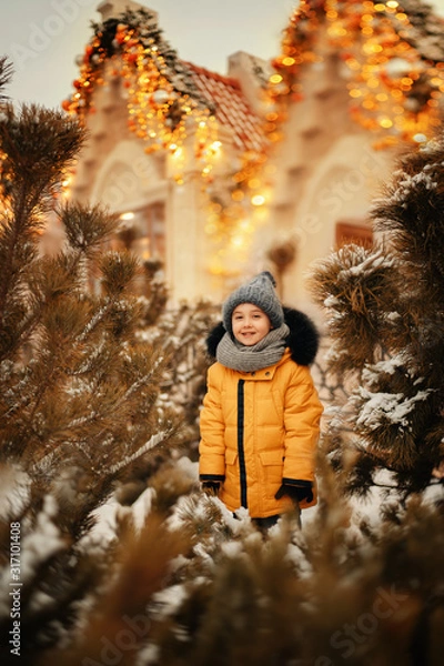 Obraz cute boy standing between pine trees on the street decorated with christmas lights at winter time