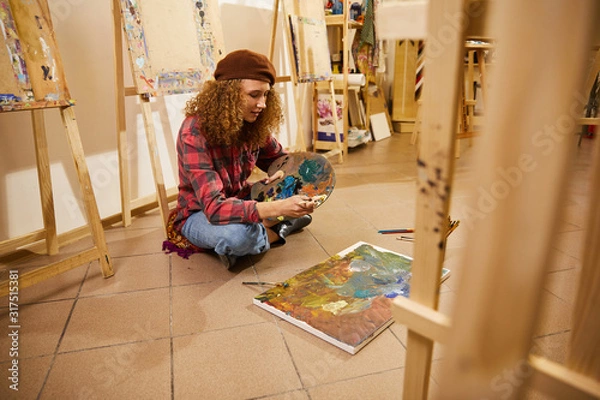 Fototapeta Curly girl sits on a floor and draw a painting with oils
