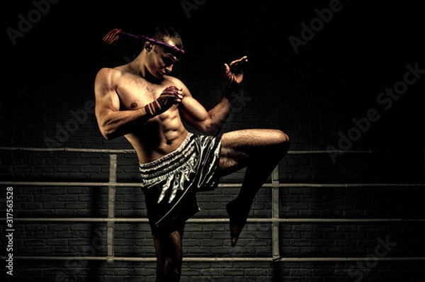 Obraz Kickboxer in the ring surrounded by searchlights stretches before the fight. Makes swing movements with his knee. Mongkhon.