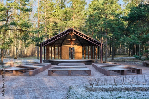 Fototapeta Wooden house stage for free musicians open air performances in pine forest in sunlight, in front of stage many benches covered with snow. Raising  culture of people, music for all, art theater in park