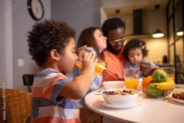 Fototapeta Happy ethnic family with two children having Breakfast sitting at the dining table in the kitchen hugging each other. Son in the foreground drinking juice from a glass