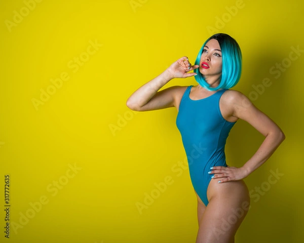 Fototapeta Beautiful girl in a blue wig and a bright bikini posing on a yellow background. Woman with artificial hair and a swimsuit emphasizing the buttocks.