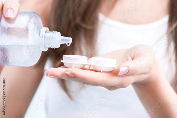 Fototapeta Closeup female hands are holding container for optical soft lens. Young woman is pouring contact lens solution liquid for disinfecting and clearing. Ophthalmic diseases and treatment concept.