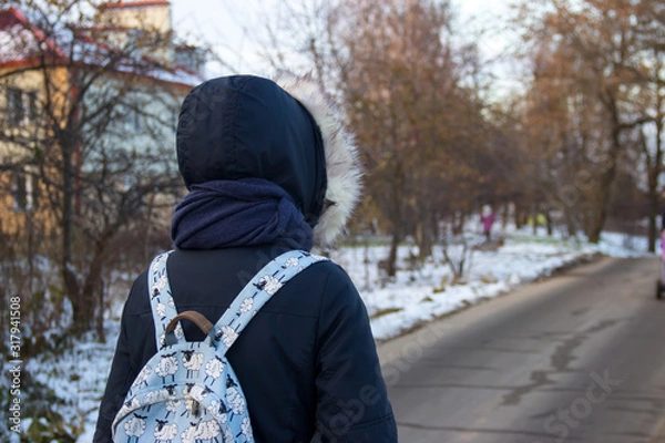 Obraz The girl in a blue jacket with a hood, scarf and blue backpack walking along the road, looking into the distance. End of autumn, early winter. On the side of the road is snow and trees