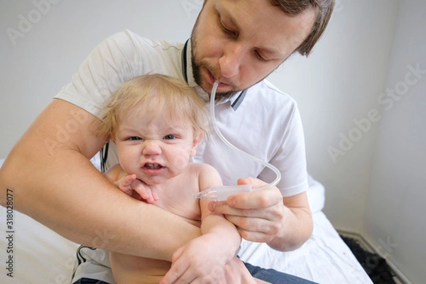 Fototapeta in the white room, a baby with snot sits on dad’s lap. Dad and son are treating and dad is cleaning his son’s nose with snot