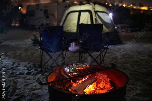 Obraz Cooking Aluminum Foil Wrapped Food on a Camp Fire Ring Grill with Tent in the Background