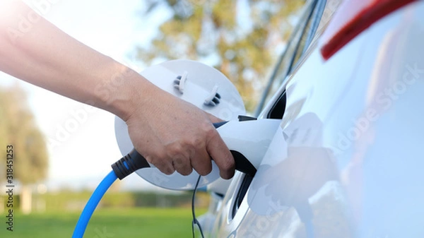 Fototapeta Electric car or ev is charging at station . man use the white power cable and plug on nature background. eco and clean energy concept with the mobile phone smart technology. zero gas no transmission