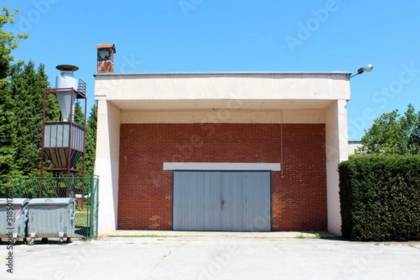 Fototapeta Large red bricks garage with elongated garage doors made from narrow wooden boards with faded color next to small storage silo surrounded with dense hedge and trees on clear blue sky background