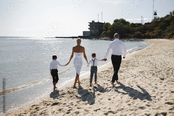Fototapeta Back view of parents and children are holding hands together and walking on the beach on the sunny summer day, dressed in white stylish clothes