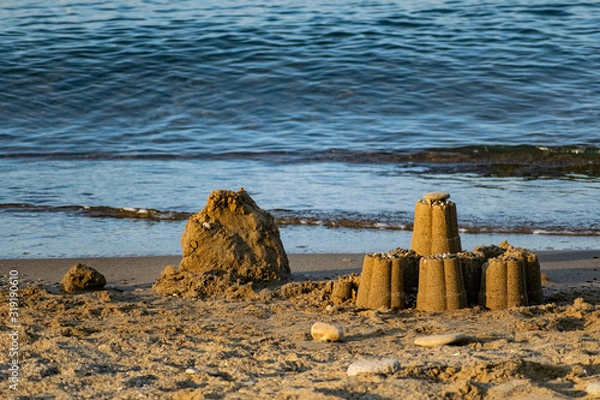 Fototapeta  Children's sand figurines on the beach. Buildings made of water, sand and stones, illuminated by sunlight from the side, at sunset, a beautiful photo for memory