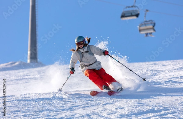 Obraz Girl On the Ski. a skier in a bright suit and outfit with long pigtails on her head rides on the track with swirls of fresh snow. Active winter holidays, skiing downhill in sunny day. Woman skier