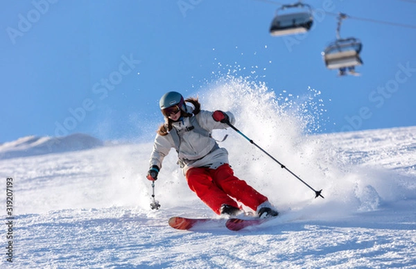 Obraz Girl On the Ski. a skier in a bright suit and outfit with long pigtails on her head rides on the track with swirls of fresh snow. Active winter holidays, skiing downhill in sunny day. Woman skier