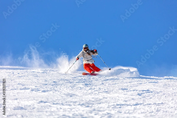 Obraz Girl On the Ski. a skier in a bright suit and outfit with long pigtails on her head rides on the track with swirls of fresh snow. Active winter holidays, skiing downhill in sunny day. Woman skier
