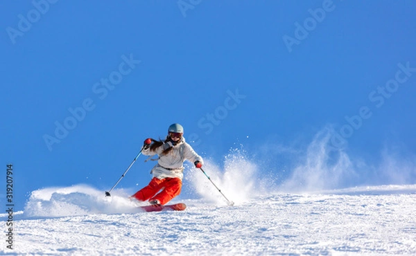 Obraz Girl On the Ski. a skier in a bright suit and outfit with long pigtails on her head rides on the track with swirls of fresh snow. Active winter holidays, skiing downhill in sunny day. Woman skier