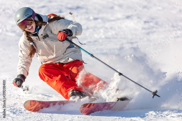 Obraz Girl On the Ski. a skier in a bright suit and outfit with long pigtails on her head rides on the track with swirls of fresh snow. Active winter holidays, skiing downhill in sunny day. Woman skier